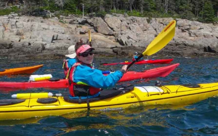 a person in a yellow kayak smiles at the camera as they paddle along a rocky shoreline in maine